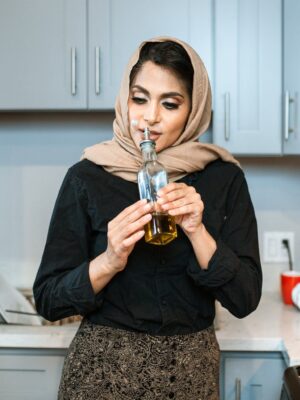 Positive stylish ethnic female in headscarf smelling aromatic olive oil in bottle while cooking dinner in contemporary light kitchen