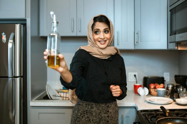 Smiling Arabic female in stylish wear and headscarf demonstrating glass bottle of olive or sunflower oil while cooking dinner in modern light kitchen
